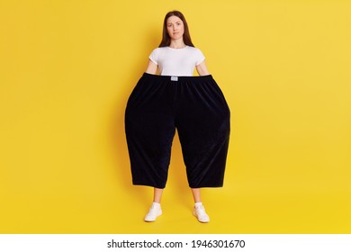 Full Length Portrait Of Puzzled Person Wearing Too Big Pants And White T Shirt Isolated On Yellow Color Background, Lady Frowning Face, Keeps Hands In Trousers.