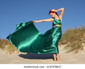 Full length portrait of  red haired woman wearing a  beautiful  long green  silk toga gown. Standing  pose with gestural hands at  ocean beach landscape background. - Shutterstock ID 2137478617