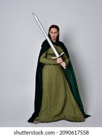 Full length portrait of red head girl wearing  green Celtic medieval gown and velvet cloak. Standing  pose holding a sword weapon.  isolated on  studio background 