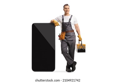 Full length portrait of a repairman in a uniform holding a tool box and leaning on a big smartphone isolated on white background - Shutterstock ID 2104921742