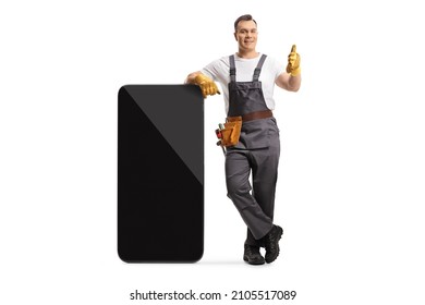 Full length portrait of a repairman in a uniform leaning on a big smartphone and gesturing a thumb up sign isolated on white background - Shutterstock ID 2105517089
