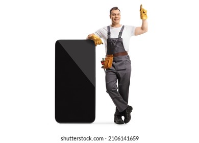 Full length portrait of a repairman in a uniform leaning on a big smartphone and pointing up isolated on white background - Shutterstock ID 2106141659