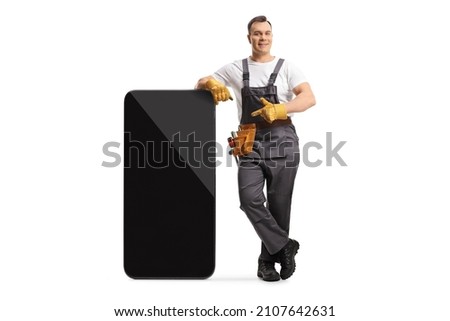 Full length portrait of a repairman in a uniform leaning on a big mobile phone and pointing at the screen isolated on white background