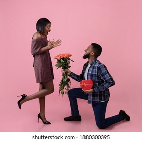 Full Length Portrait Of Romantic Black Guy Giving His Girlfriend Flowers And Gift For Valentine's Day Over Pink Studio Background. Excited Woman Receiving Roses And Present From Her Boyfriend