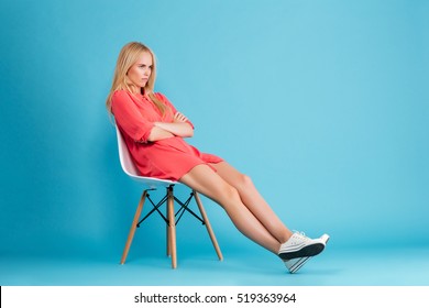 Full Length Portrait Of A Sad Grumpy Girl In Red Dress Sitting On Chair And Looking Away Isolated On The Blue Background