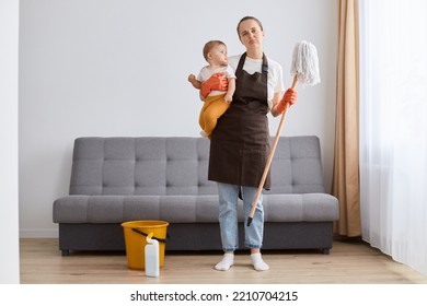 Full Length Portrait Of Sad Upset Woman Wearing Apron Washing Floor With Mop At Home, Holding Her Infant Baby In Hands, Combines Maternity Leave And Household Chores, Looking At Camera With Pout Lips.