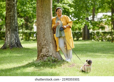 Full Length Portrait Of Senior Black Woman Leaning On Tree In Park While Walking Cute Dog And Drinking Coffee, Copy Space