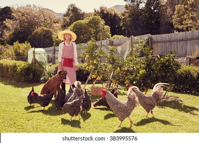 Full Length Portrait Of A Senior Woman Standing Happily In Her Backyard, With Her Dog Sitting Beside Her And A Group Of Healthy Free Range Chickens On The Grass In Front Of Her