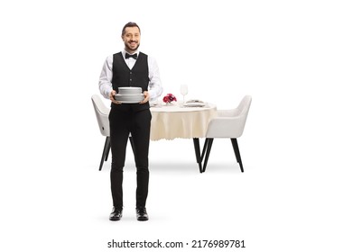 Full length portrait of a server holding a pile of plates and standing in front of a table isolated on white background