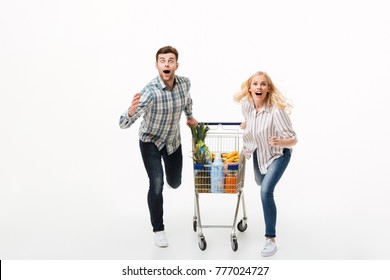 Full length portrait of a shocked couple running with a supermarket trolley isolated over white background