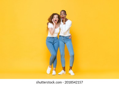 Full length portrait of shocked interracial millennial woman friends gasping with hands cupping mouths in isolated studio yellow color background