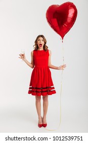 Full Length Portrait Of A Shocked Young Woman Dressed In Red Dress Holding Air Balloon While Drinking Glass Of Champagne And Celebrating Isolated Over White Background