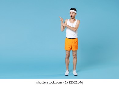 Full Length Portrait Shocked Young Fitness Man With Skinny Body Sportsman In White Headband Shirt Shorts Pointing Index Fingers Aside Isolated On Blue Background. Workout Gym Sport Motivation Concept