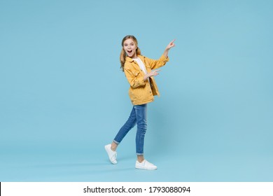 Full Length Portrait Side View Of Excited Little Kid Girl 12-13 Years Old In Yellow Jacket Isolated On Blue Background. Childhood Lifestyle Concept. Mock Up Copy Space. Pointing Index Fingers Aside