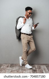 Full Length Portrait Of A Smiling African Man In Sunglasses Holding Jacket Over Shoulder And Typing Message On Mobile Phone Isolated Over Gray Background