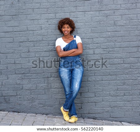 Full length portrait of smiling african woman in dungarees standing against gray wall