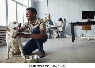 Full length portrait of smiling African-American man petting dog while working in office, pet friendly workspace, copy space - Powered by Shutterstock