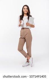 Full length portrait of a smiling asian businesswoman carrying laptop computer and cup of coffee to go while standing isolated over white background