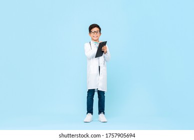 Full Length Portrait Of Smiling Asian Child Aspiring To Be Future Doctor Writing Prescription In Clipboard In Blue Isolated Background