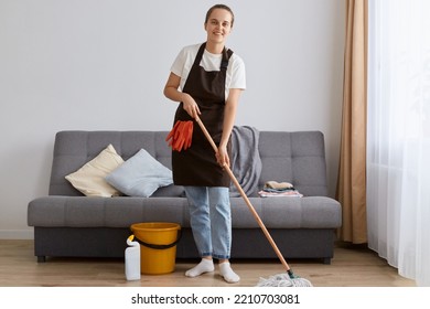 Full Length Portrait Of Smiling Attractive Woman Wearing Jeans And Brown Apron, Washing Floor With Mop At Home, Looking At Camera With Happy Positive Emotions, Likes To Do Work About The House.