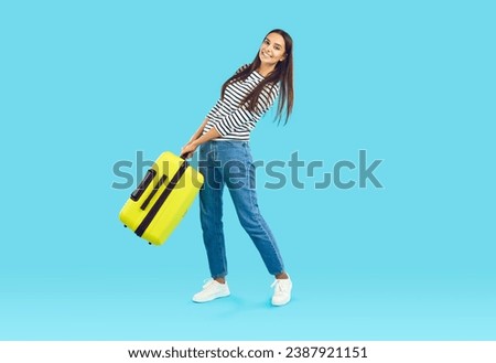 Full length portrait of smiling brunette cheerful girl carrying heavy yellow suitcase in hands on blue background. She is wearing jeans and striped blouse. Travelling, tourism, journey, trip concept.