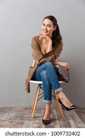 Full length portrait of a smiling casual asian woman sitting on a chair and looking at camera over gray background