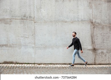 Full Length Portrait Of A Smiling Casual Man Walking On A City Street Against Gray Wall