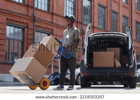 Full length portrait of smiling delivery worker unloading boxes outdoors in sunlight, copy space