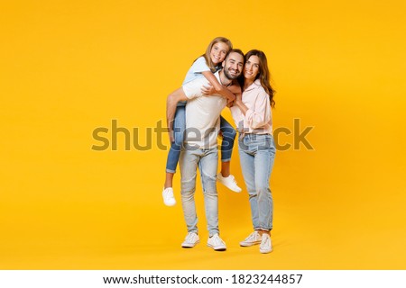 Full length portrait of smiling funny young parents mom dad with child kid daughter teen girl in t-shirts giving piggyback ride to joyful sit on back isolated on yellow background. Family day concept