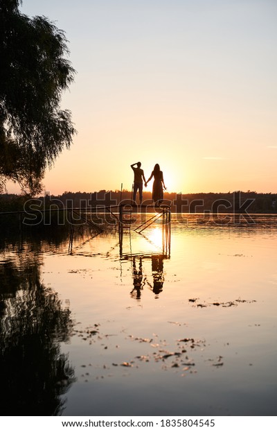 Full length portrait of smiling hipster man\
and his beloved lady holding hands while staying at the wooden\
bridge near the water and looking at the\
sunset
