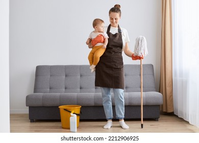 Full Length Portrait Of Smiling Joyful Woman In Apron Washing Floor With Mop At Home, Doing Domestic Chores, Cleaning Apartment, Holding Infant Baby In Hands.