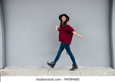 Full length portrait of a smiling joyful asian girl dressed in hat and sweater holding coffee cup while walking on a city street