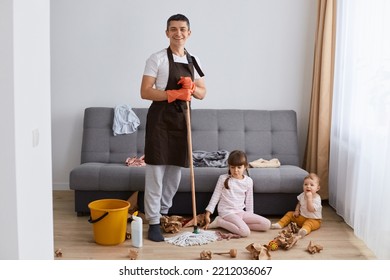 Full Length Portrait Of Smiling Positive Caucasian Man Wearing Brown Apron Washing Floor With Mop At Home, His Kids Playing In Living Room In Mess, Guy Looking At Camera With Happiness.