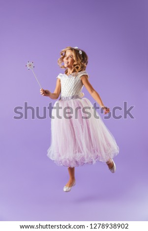 Full length portrait of a smiling pretty girl dressed in a princess dress isolated over violet background, holding magic wand, jumping