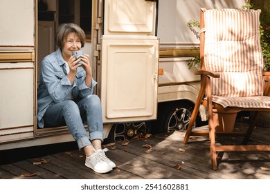 Full length portrait of smiling relaxed old senior elderly traveler woman grandmother drinking coffee resting on the porch of trailer camper van motor wheel home - Powered by Shutterstock