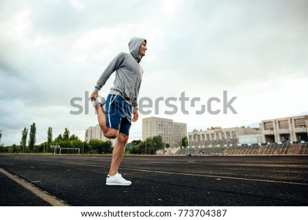 Full length portrait of a smiling sportsman doing exercises for legs, with headphones, at the stadium