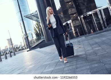Full length portrait of a smiling successful businesswoman pulling suitcase and talking on the phone in the city.