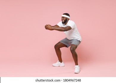 Full Length Portrait Of Smiling Tired Young Bearded African American Fitness Sports Man 20s In Headband T-shirt Doing Exercise Squatting Spending Time In Gym Isolated On Pink Color Background Studio