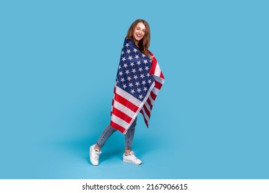 Full length portrait of smiling woman wearing striped sweater, standing wrapped in American flag, celebrating labor day, government employment support. Indoor studio shot isolated on blue background.