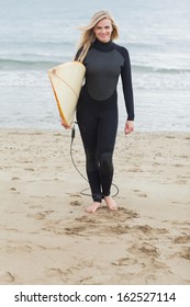 Full Length Portrait Of A Smiling Young Woman In Wet Suit Holding Surfboard At Beach