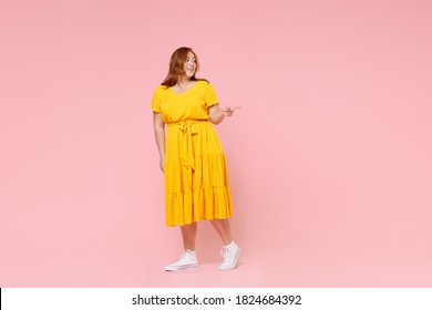 Full Length Portrait Smiling Young Redhead Plus Size Body Positive Chubby Overweight Woman In Yellow Dress Pointing Index Finger Aside On Mock Up Copy Space Isolated On Pastel Pink Color Background