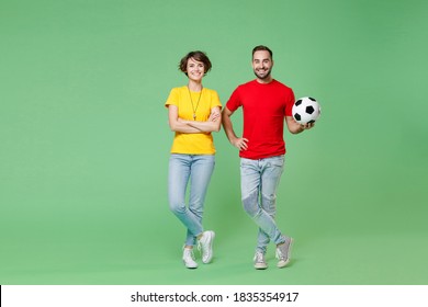 Full Length Portrait Of Smiling Young Couple Friends Sport Family Woman Man Football Fans In Yellow Red T-shirts Cheer Up Support Favorite Team With Soccer Ball Isolated On Green Background Studio