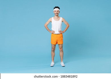 Full Length Portrait Smiling Young Fitness Man With Skinny Body Sportsman In Headband Shirt Shorts Standing With Arms Akimbo On Waist Isolated On Blue Background. Workout Gym Sport Motivation Concept