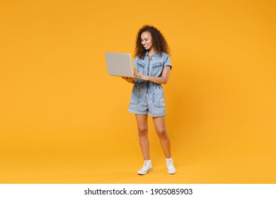 Full length portrait of smiling young african american woman 20s in denim clothes isolated on yellow wall background studio portrait. People lifestyle concept. Working on laptop pc computer at home