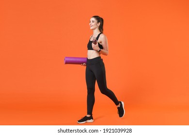 Full length portrait of smiling young fitness sporty woman in black sportswear posing going training working out hold bottle of water yoga mat looking aside isolated on orange color background studio