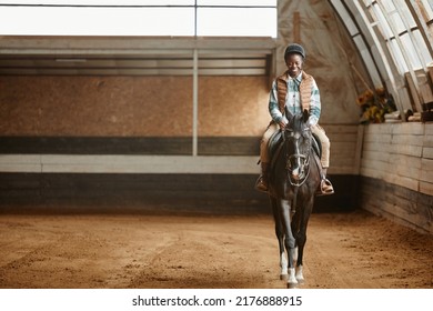 Full length portrait of smiling young woman riding horse in indoor arena at horse ranch or practice stadium, copy space