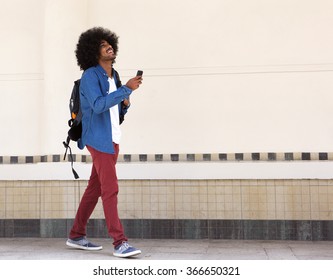 Full Length Portrait Of A Smiling Young Black Man Walking With Bag And Mobile Phone