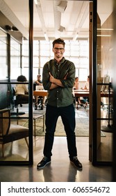 Full Length Portrait Of Smiling Young Man Standing In Doorway Of Office With His Arms Crossed. Creative Male Executive At Startup With People Working In Background.