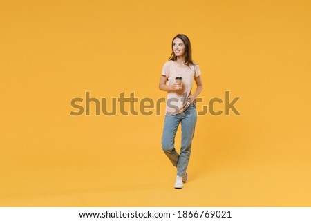 Full length portrait of smiling young brunette woman 20s wearing pastel pink casual t-shirt posing hold paper cup of coffee or tea looking aside isolated on bright yellow color wall background studio