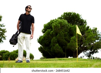 Full length portrait of stylish golfer man in glasses standing on beautiful golf course looking away, handsome brunette hair man with golf bag on shoulder and hand in pocket standing on golf course
